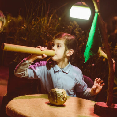 A When the World Turns production photo. A child holds up a cardboard tube, using it to make sounds. An adult leans towards the tube, listening. They sit at a small table in a darkened space, surrounded by plants. They are illuminated by a lamp on the table. Photographer: Theresa Harrison