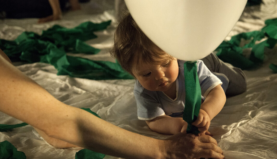 A How High The Sky production photo. A close up of a baby lying on a soft white surface, holding the string of a white balloon and a green streamer. An adult's arm is stretched towards them. They are illuminated with warm theatre lighting.