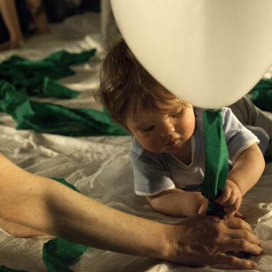 A How High The Sky production photo. A close up of a baby lying on a soft white surface, holding the string of a white balloon and a green streamer. An adult's arm is stretched towards them. They are illuminated with warm theatre lighting.