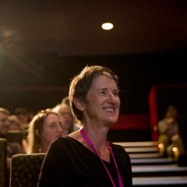Sue Giles at a 2017 Polyglot Theatre 5678 Film Club event. She is seated in a cinema, looking at someone out of frame, smiling. Other people are visible behind her. Photographer: Theresa Harrison