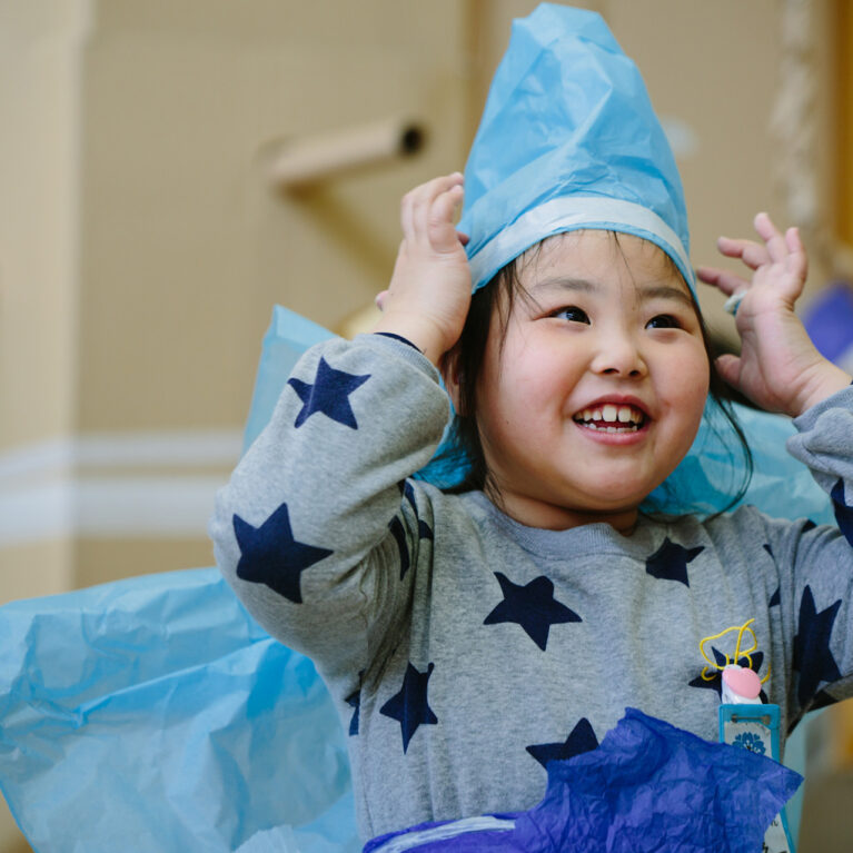 A Paper Planet production photo. A child in a grey jumper with a blue star pattern and a handmade blue tissue paper costume smiles at someone out of frame. Their hands are raised to their paper hat, and they are surrounded by tall brown cardboard trees.