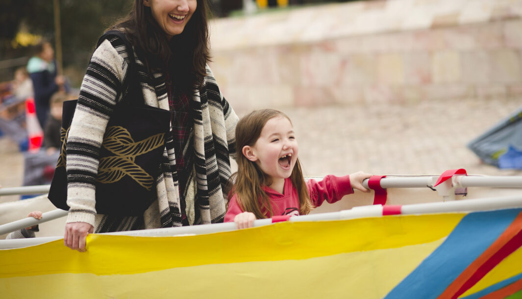 A Boats production photo. An adult and child stand in a yellow and blue vessel, holding it around themselves. They are both smiling widely.