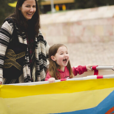 A Boats production photo. An adult and child stand in a yellow and blue vessel, holding it around themselves. They are both smiling widely.