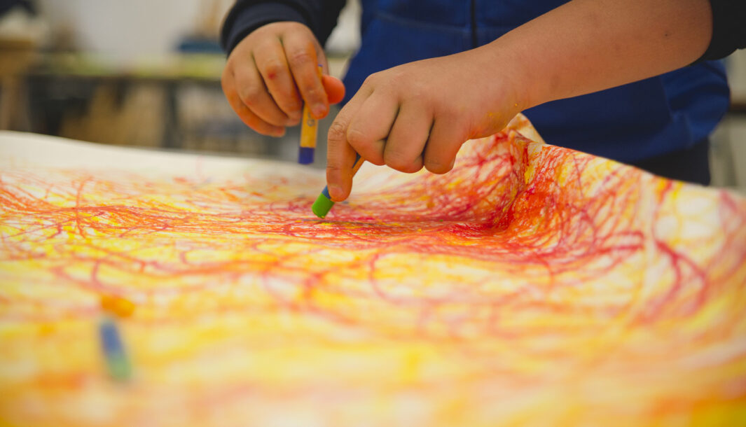 A school residency photo. A close up of a child's hands, holding green and blue pastels, about to drawn on paper covered in orange and yellow scribbles and swirls.