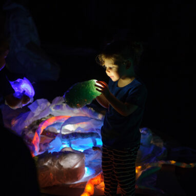A Light Pickers creative development photo. A small child stands in a darkened space, next to a pile of colourful, abstract glowing objects. They hold a glowing object close to their face, looking at it intently.