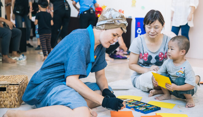A Cerita Anak (Child's Story) production photo. A Polyglot artist sits with an adult and child on the floor, creating colourful sea creatures with construction paper.