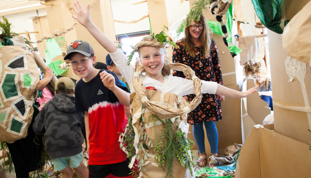 A Paper Planet production photo. A child wears a paper branch wrapped around their shoulders and arms, smiling with their arms outstretched. Another child stands to their right, smiling. Behind them are paper decorations and trees made from cardboard. Photo: Mark Gambino.