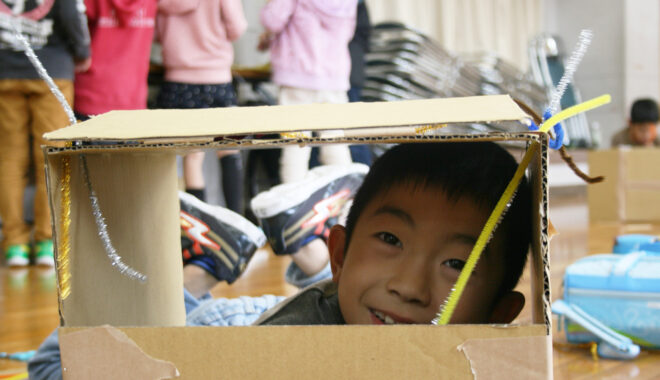 A workshop photo. A child peers out of a cardboard box, smiling at the camera.