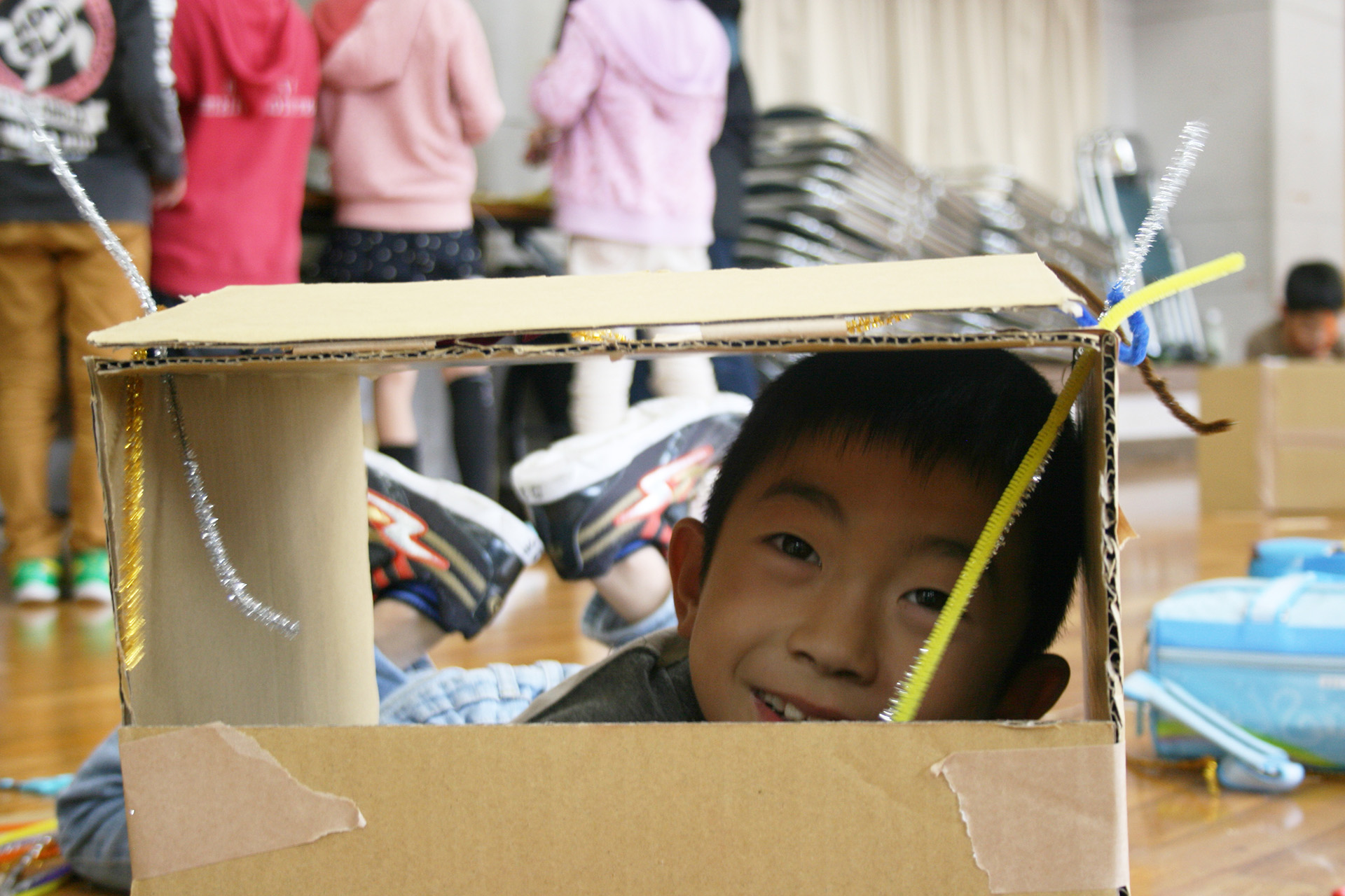 A workshop photo. A child peers out of a cardboard box, smiling at the camera.