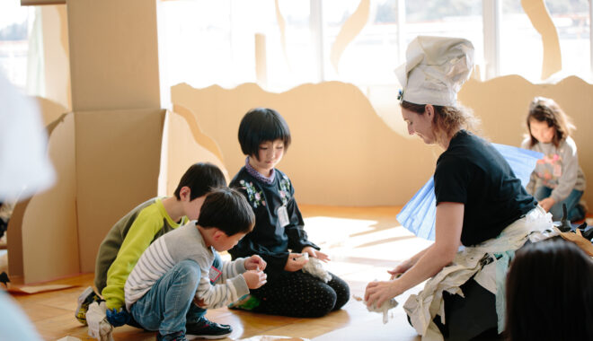 A Paper Planet workshop photo. A Polyglot artist wearing a handmade paper costume kneels on the floor with three children, making paper creations. They are surrounded by tall brown cardboard trees.