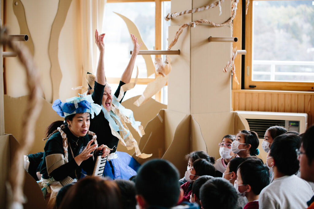 Paper Planet workshop photo: Ai Ueda. Two excited artists sit in front of a group of children, talking and gesturing to them. they are surrounded by tall brown cardboard trees.