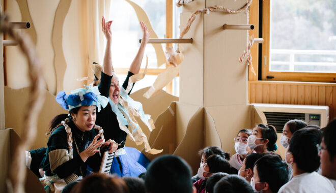 A Paper Planet workshop photo. Two excited artists sit in front of a group of children, talking and gesturing to them. they are surrounded by tall brown cardboard trees.
