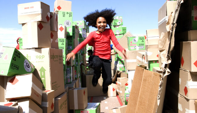 A We Built This City production photo. A child with dark curly hair, wearing a red shirt, leaps into the air, smiling. They are surrounded by hundreds of cardboard boxes piled high.