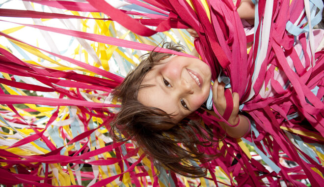 A Tangle production photo. A child with short dark hair is suspended upside down amongst hundreds of strands of colourful elastic. They are smiling at the camera.