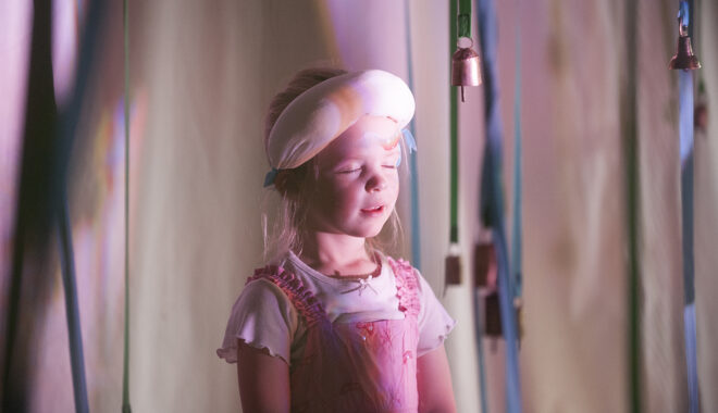 A Bellbird production photo. A child in a pink dress stands amongst suspended blue and green ribbons with small bells attached to the ends. Their eyes are closed and a white blindfold is pushed up onto their forehead.
