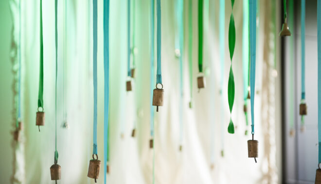 A Bellbird production photo. Green and blue ribbons hang in front of a white cloth. Each ribbon has a small gold bell attached to the end.