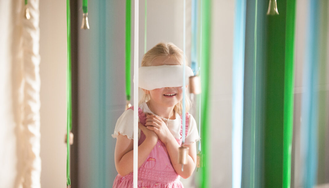 A Bellbird production photo. A child in a pink dress, with a large white blindfold over their eyes, stands amongst suspended blue and green ribbons with bells tied to the ends. The child's hands are clasped and they are smiling.