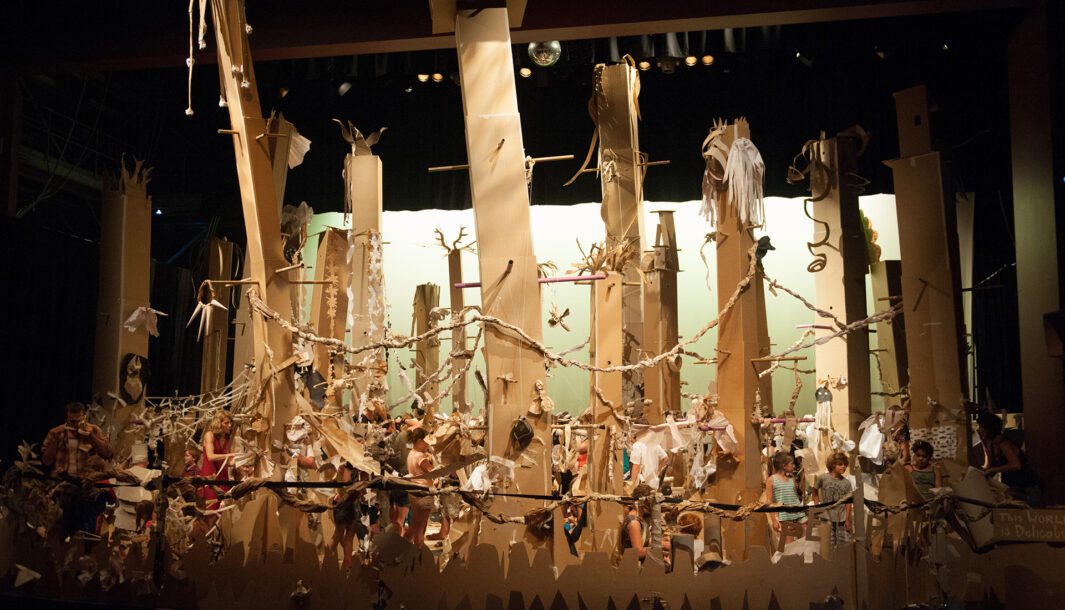 A Paper Planet production photo. A wide shot showing a forest of tall, brown cardboard trees in a darkened theatre space. The trees are illuminated with theatre lighting.