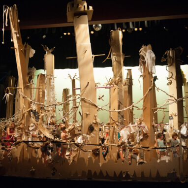 A Paper Planet production photo. A wide shot showing a forest of tall, brown cardboard trees in a darkened theatre space. The trees are illuminated with theatre lighting.