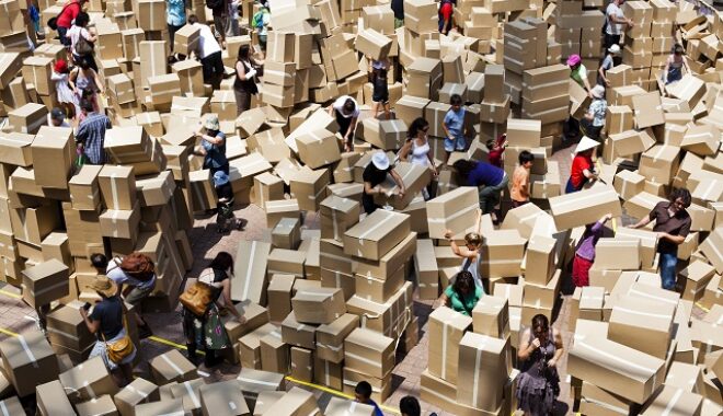 A We Built This City Production Photo. Cardboard boxes dwarf people walking beside them. People stack cardboard boxes in different heights and directions.