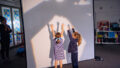 A Shadow Tricks workshop photo. Two students stand behind a tall screen in a classroom, their arms raised. A shadow shape is projected around them. A Polyglot artist watches from the other side of the screen.