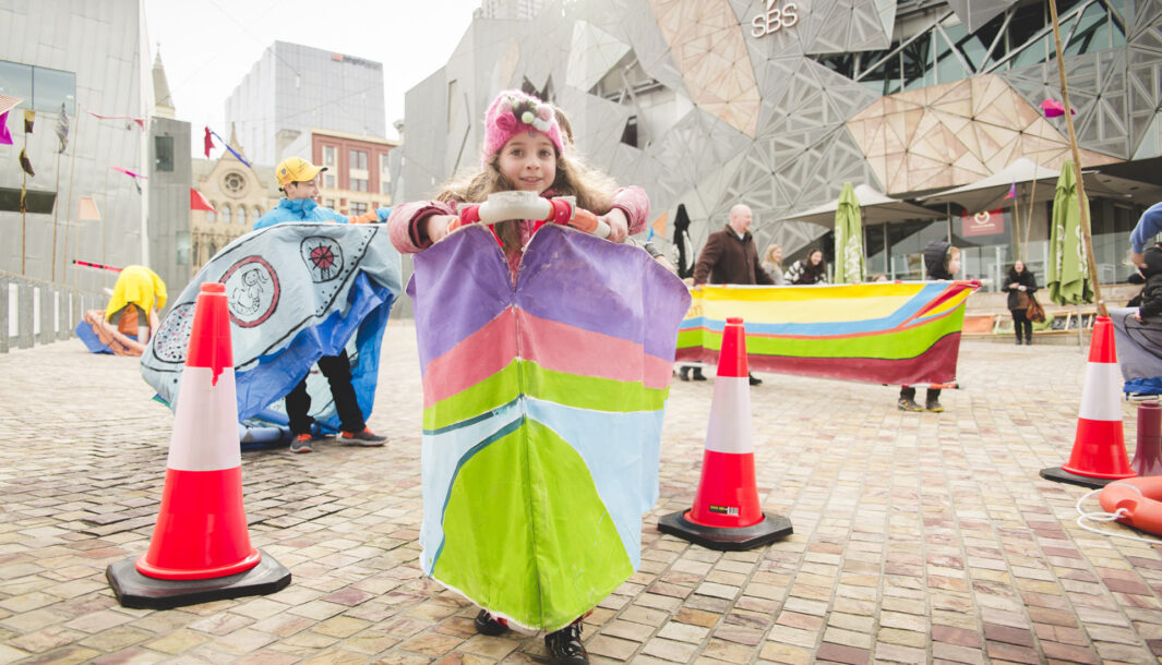 A Boats production photo. A child stands in a colourful vessel, smiling directly at the camera. Around them are other children and families in colourful vessels, and orange traffic cones.