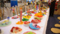 A Feast production photo. A long table covered in a white cloth is filled with colourful plates of food made from paper and cellophane.