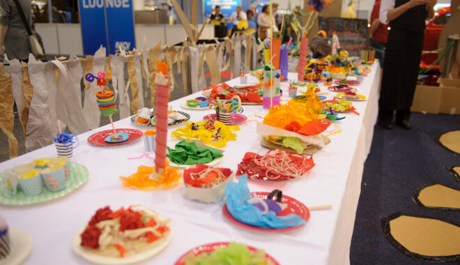 A Feast production photo. A long table covered in a white cloth is filled with colourful plates of food made from paper and cellophane.