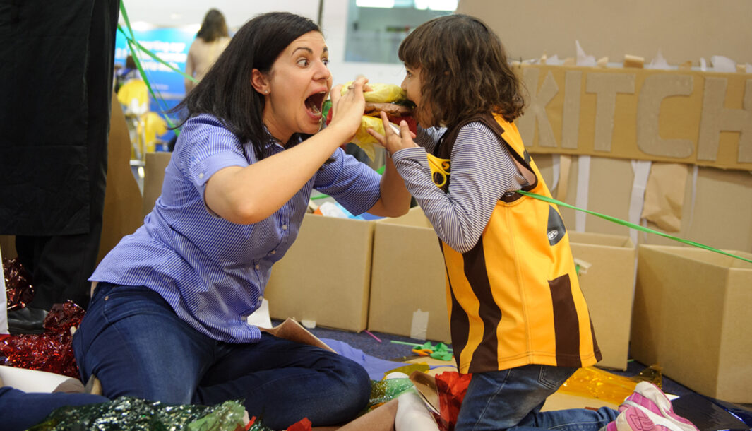 A Feast production photo. An adult and child hold up a 'burger' made from paper and cellophane between them, pretending to take enormous bits. A cardboard sign saying 'kitchen' is partially visible behind them.