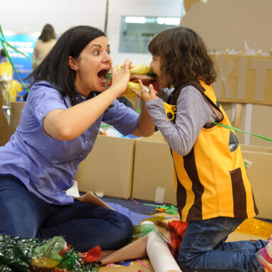 A Feast production photo. An adult and child hold up a 'burger' made from paper and cellophane between them, pretending to take enormous bits. A cardboard sign saying 'kitchen' is partially visible behind them.