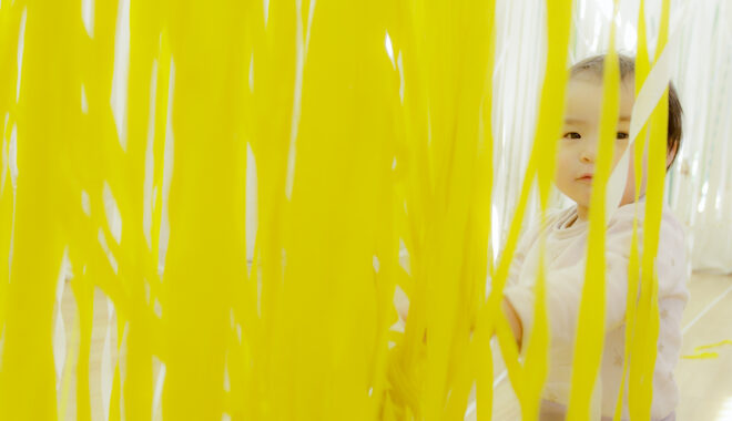 A Bellbird production photo. A baby sits behind hundreds of strands of suspended yellow and white ribbons.