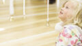A Bellbird production photo. A child with fair hair sits on a wooden floor, gazing upwards, fascinated. White ribbons with small gold bells attached to the ends are suspended in the air around them.