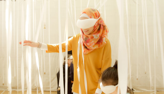 A Bellbird production photo. An adult and a child, both wearing white blindfolds, hold hands as they move through hundreds of white ribbons suspended in the air.
