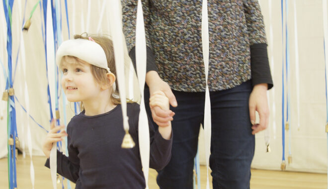 A Bellbird production photo. A child with a white blindfold pushed up onto their forehead looks around the space smiling, holding their adults hand. They are surrounded by suspended blue and white ribbons with small gold bells attached to the ends.