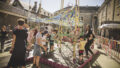 A Tangle production photo. Tall golden poles stand in a courtyard surrounded by heritage buildings. Hundreds of strands of colourful elastic are woven amongst them, and children and families play throughout.
