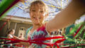 A Tangle production photo. A child wearing a tie-dye shirt smiles and reaches over the camera, their long fair over their face. They are surrounded by hundreds of colourful strands of elastic.