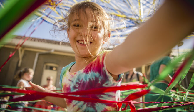 A Tangle production photo. A child wearing a tie-dye shirt smiles and reaches over the camera, their long fair over their face. They are surrounded by hundreds of colourful strands of elastic.