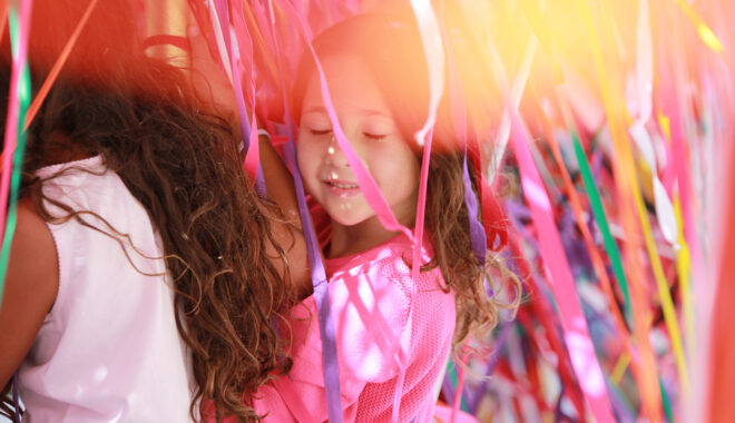 A Tangle production photo. A child stands against a mass of hundreds of strands of colourful elastic. Their eyes are closed and they are smiling.