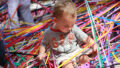 A Tangle production photo. A baby sits on a 'net' of hundreds of strands of colourful elastic, looking at it with interest.