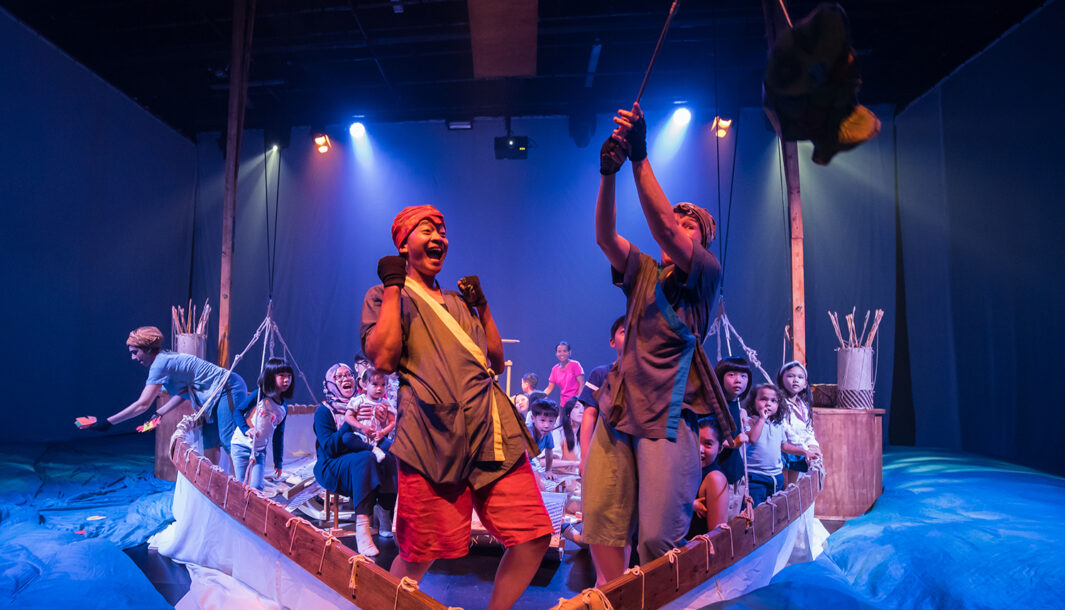 A Cerita Anak production photo. Two Polyglot artists wearing blue and red costumes stand at the bow of the boat, joyfully celebrating a special moment. Families sit in the boat behind them. They are surrounded by billowing blue silk cloth, and illuminated with theatre lighting.