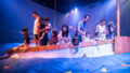 A Cerita Anak production photo. Families stand on the boat, fishing for paper sea creatures in the billowing blue silk ocean. They are illuminated with bright theatre lighting.