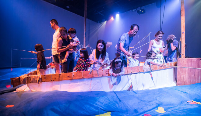 A Cerita Anak production photo. Families stand on the boat, fishing for paper sea creatures in the billowing blue silk ocean. They are illuminated with bright theatre lighting.