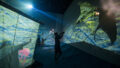 A Cerita Anak production photo. A Polyglot artist is seen in silhouette, manipulating a sea creature shadow puppet against huge screens illuminated with green and blue children's drawings.