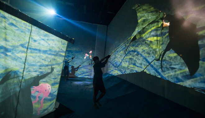 A Cerita Anak production photo. A Polyglot artist is seen in silhouette, manipulating a sea creature shadow puppet against huge screens illuminated with green and blue children's drawings.