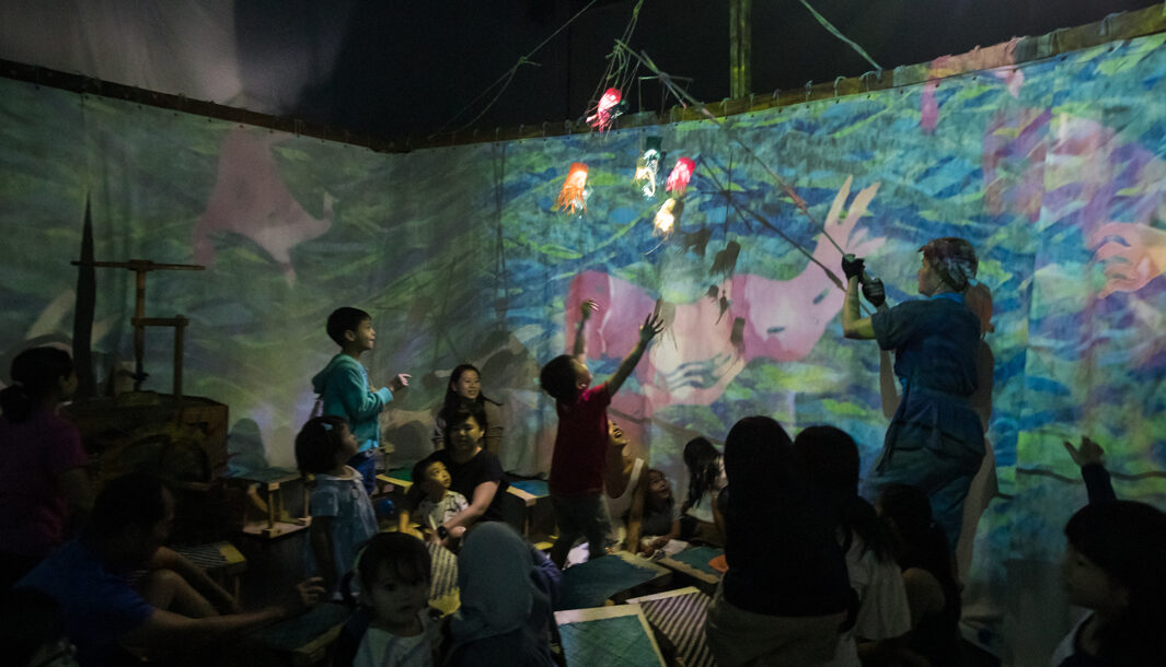 A Cerita Anak production shot. A performer holds a light display comprising of paper octopuses on a marionette in front of a crowd of children. One child has their arms outstretched towards the light display.