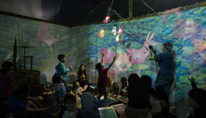 A Cerita Anak production shot. A performer holds a light display comprising of paper octopuses on a marionette in front of a crowd of children. One child has their arms outstretched towards the light display.