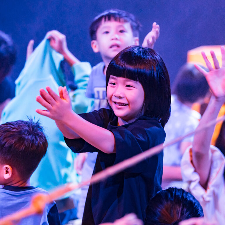 A Cerita Anak production photo. A child stands in the boat, smiling and clapping. They are surrounded by other smiling children and adults. They are illuminated by bright theatre lighting.