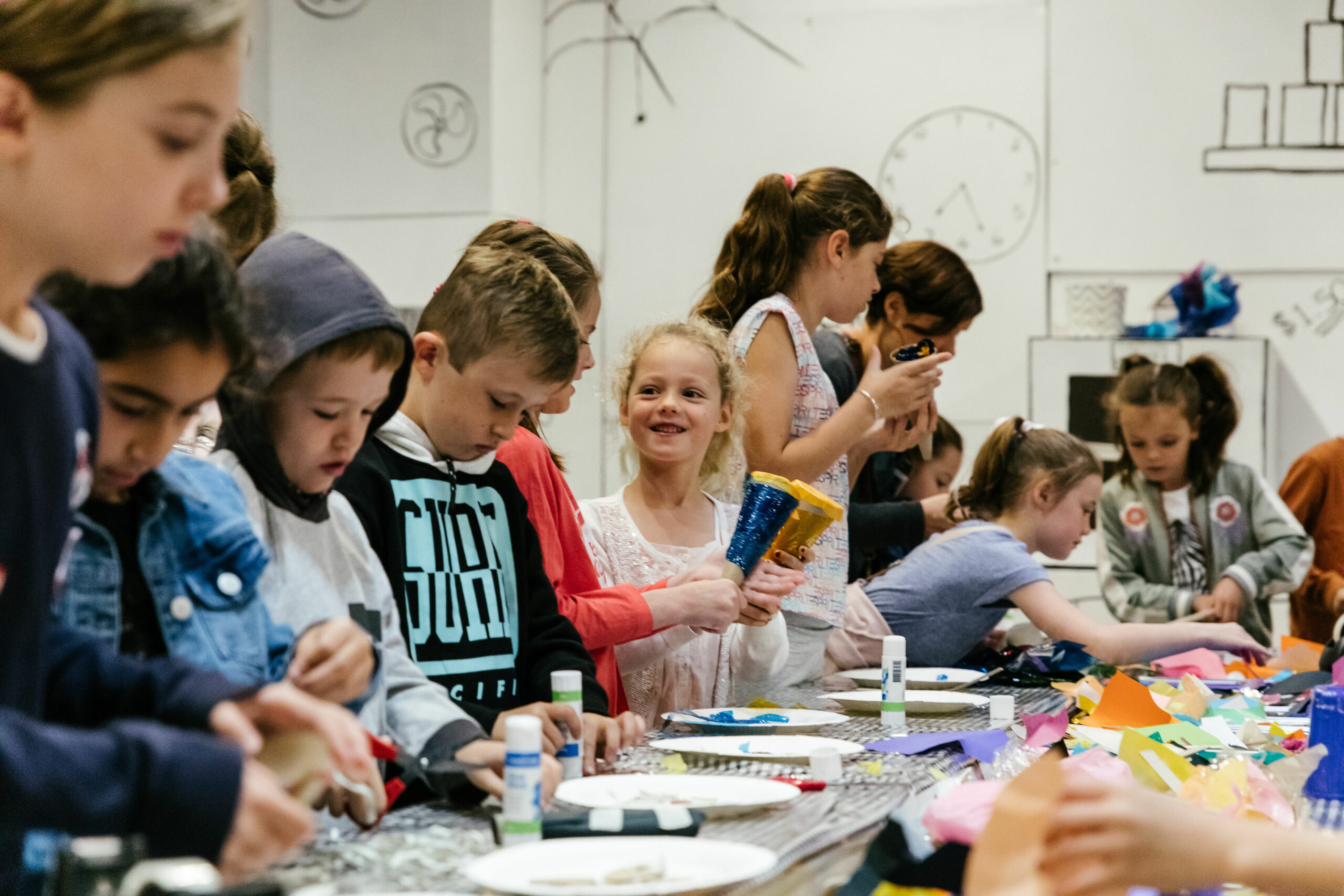 A Feast production photo. A group of children stand at a table, making food with paper, cardboard and cellophane.