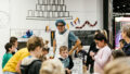 A Feast production photo. A Polyglot artist in character wearing a blue hairnet and white apron watches a group of children making paper food creations at a table. Behind them is a large white wall covered in an illustration of a tower of cans.