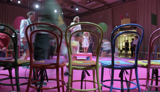 An indoor Invisible Orchestra photo. Performers in white jackets are visible behind colourful chairs. They are surrounded by running children who are motion blurs.